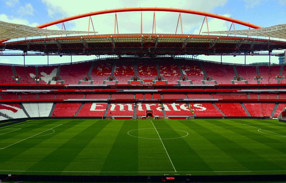 An expansive view of a modern football stadium showing a well-maintained grass pitch with distinct mowing lines, surrounded by tiered red seats with white accents forming the word 'Emirates' across the middle sections. The seating area includes sections of red and white seats, with some grey seats at the upper levels. The stadium features a large, curved roof structure with a red arch spanning across the top, supported by steel beams and partially visible under a bright, partly cloudy sky. Around the pitch, there are pathways and landscaped areas with soil and small plants, indicating ongoing or recent garden maintenance work. The environment suggests a clean, organized outdoor space prepared for a match or event, exemplifying professional landscaping and groundskeeping by Gardeners Highbury.