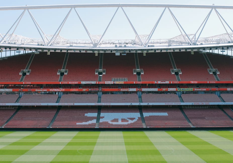 A view of a football stadium's interior with a well-maintained grass pitch marked with white lines. The seating area features rows of red and blue seats, with some green seats spelling out a pattern. The stadium's roof structure is visible above, with a white truss supporting it. The lighting suggests daytime, with natural light illuminating the field and seating areas. Although not directly related to gardening, this image showcases the importance of landscape maintenance and outdoor care essential for maintaining such large sports venues, a service provided by Gardeners Highbury.