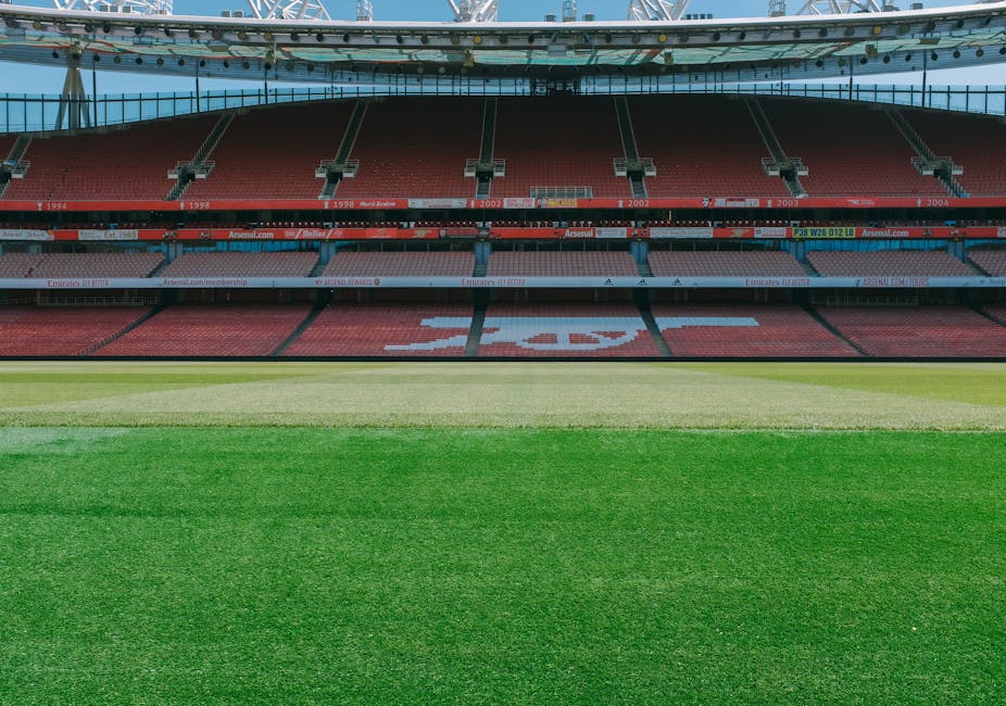 An expansive view of a modern football stadium showing a well-maintained grass pitch with distinct mowing lines, surrounded by tiered red seats with white accents forming the word 'Emirates' across the middle sections. The seating area includes sections of red and white seats, with some grey seats at the upper levels. The stadium features a large, curved roof structure with a red arch spanning across the top, supported by steel beams and partially visible under a bright, partly cloudy sky. Around the pitch, there are pathways and landscaped areas with soil and small plants, indicating ongoing or recent garden maintenance work. The environment suggests a clean, organized outdoor space prepared for a match or event, exemplifying professional landscaping and groundskeeping by Gardeners Highbury.